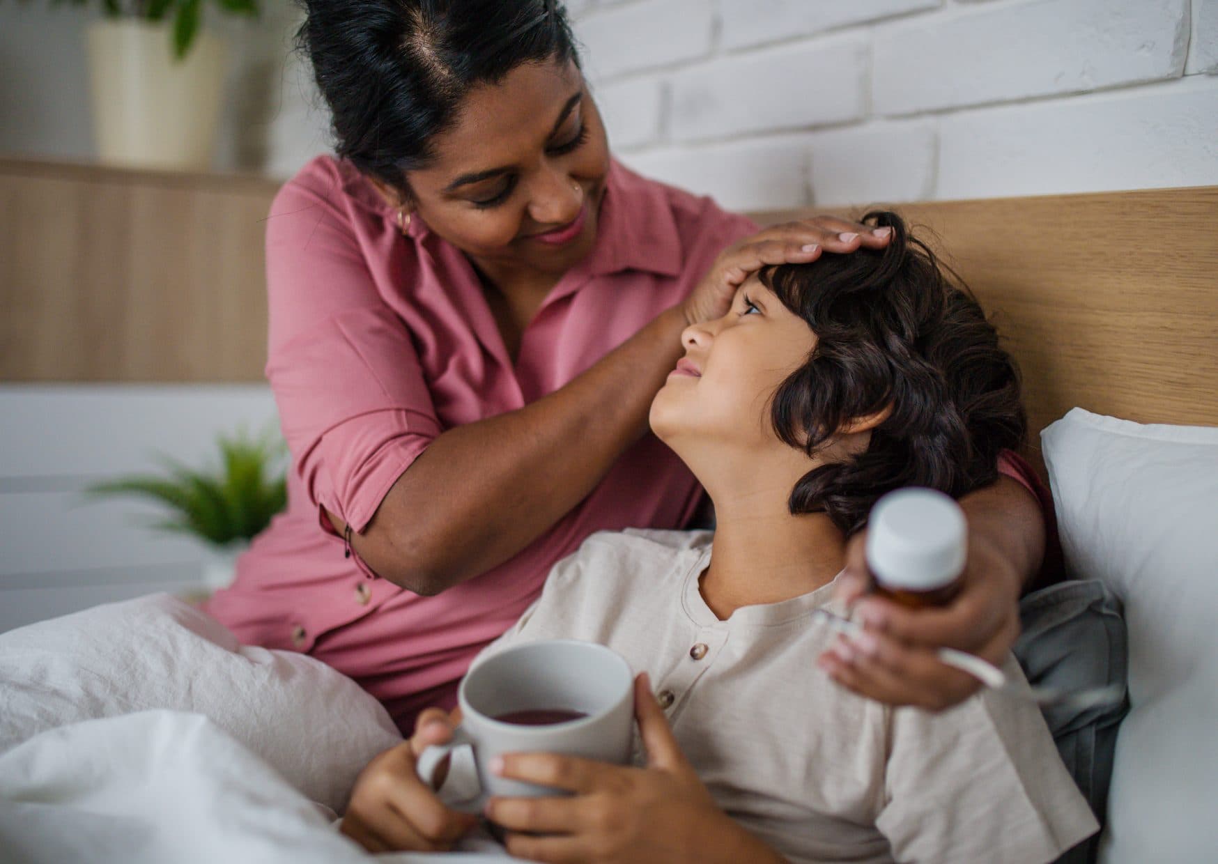 A mother sitting on a bed feeling her young son's forehead while he holds a mug and a bottle of medicine