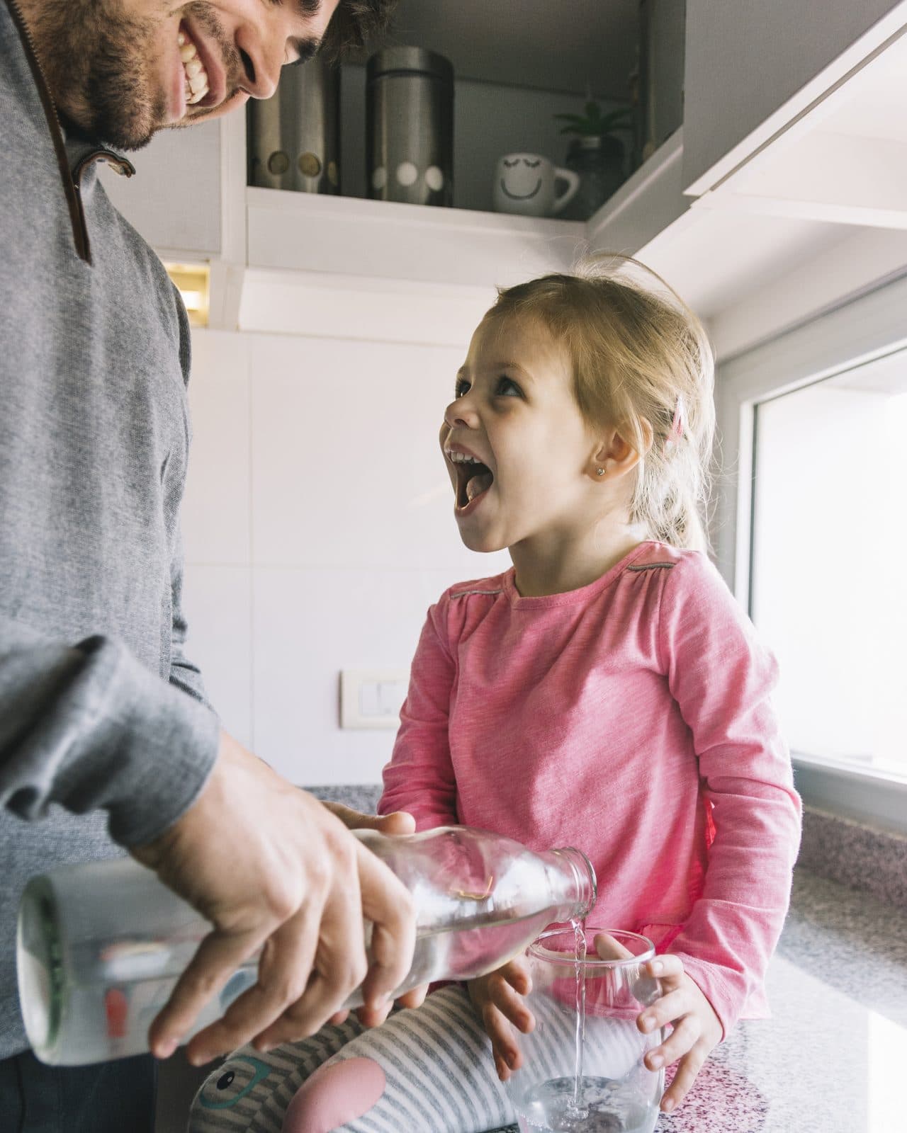 A father pouring a glass of water for his laughing young daughter in a kitchen