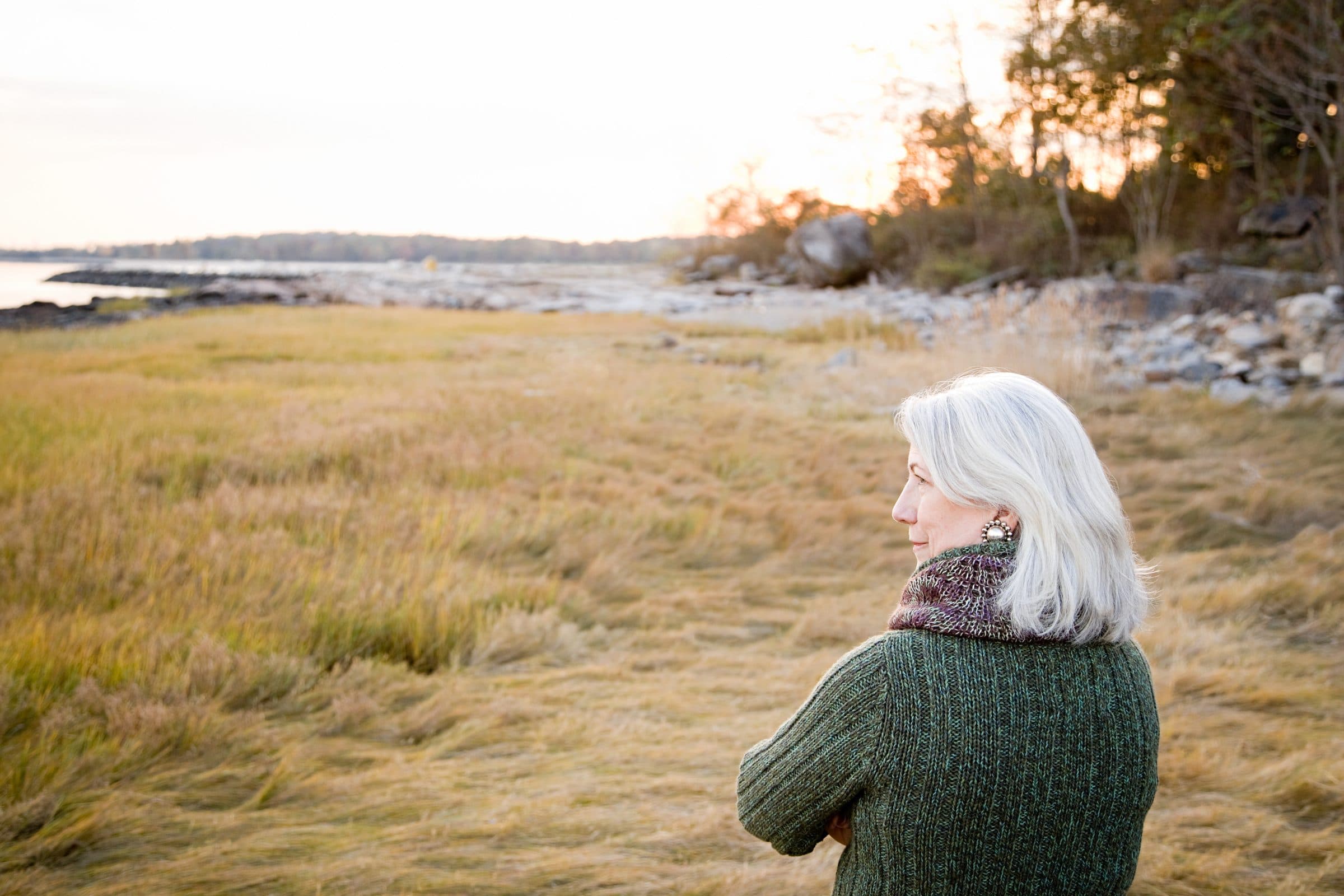 A woman with white hair in a green jumper looking out across a grassy shoreline at dusk