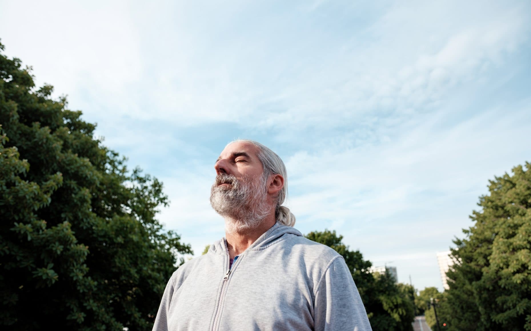 A man with grey hair and a beard standing outdoors with his eyes closed, breathing in, trees and sky behind him