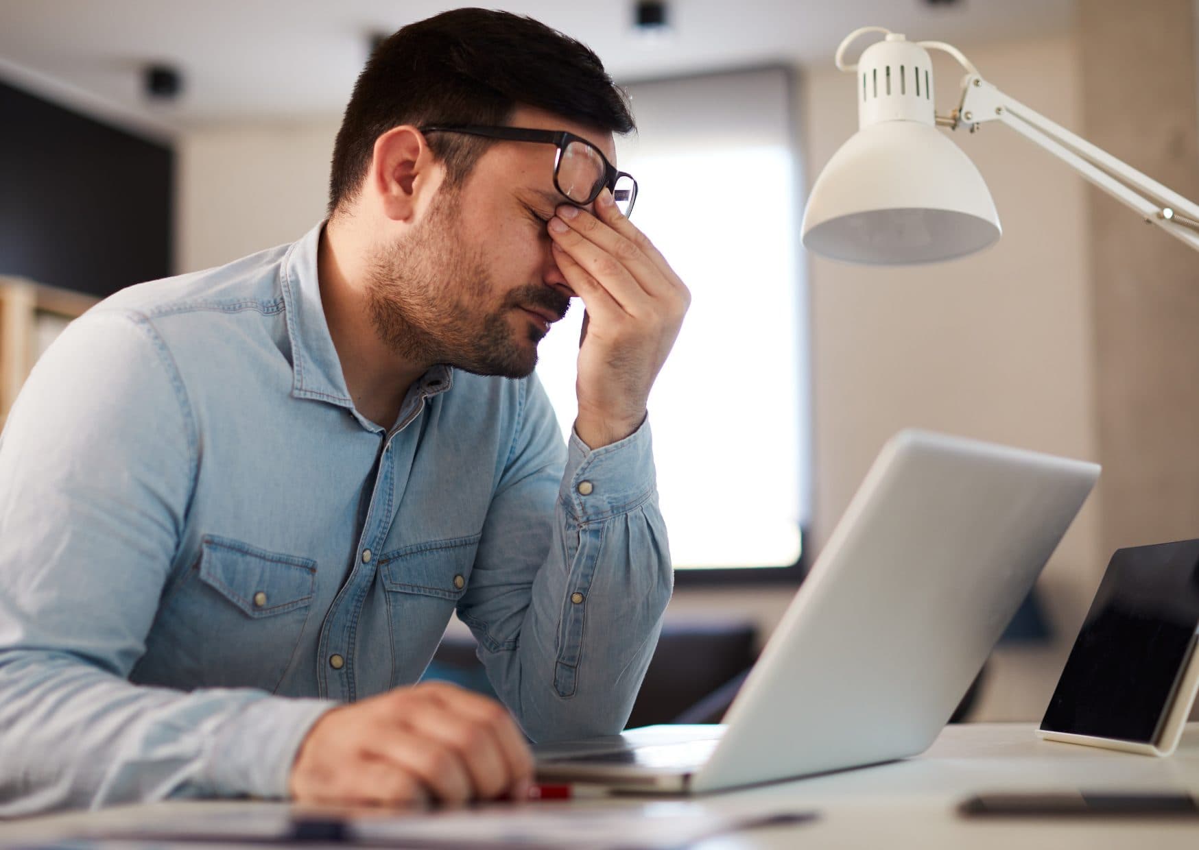 A man in glasses sitting at a laptop, pinching the bridge of his nose with his eyes closed