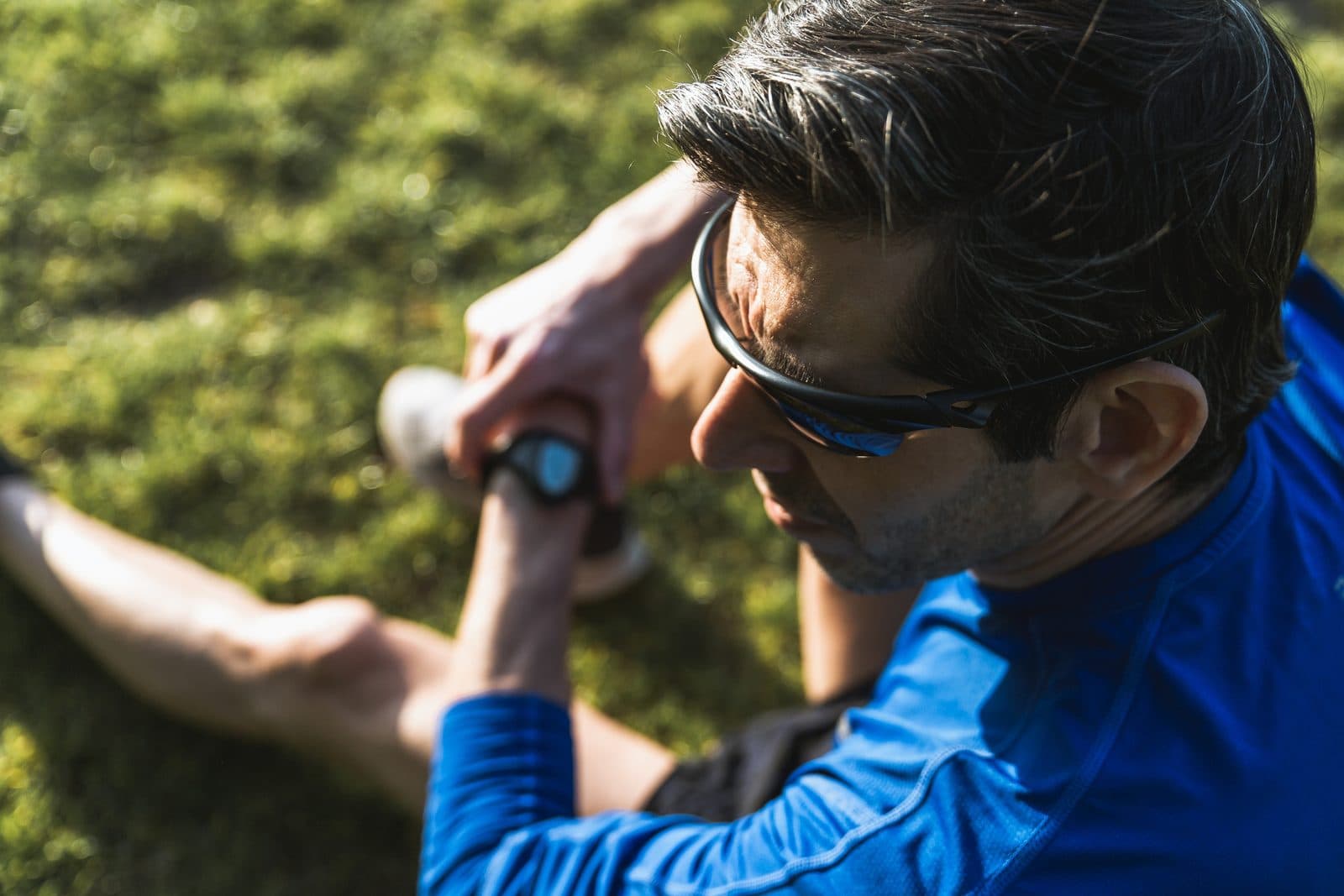 A man wearing dark sunglasses and a blue long-sleeved sports top outdoors in bright sunlight, with blurred green foliage behind him